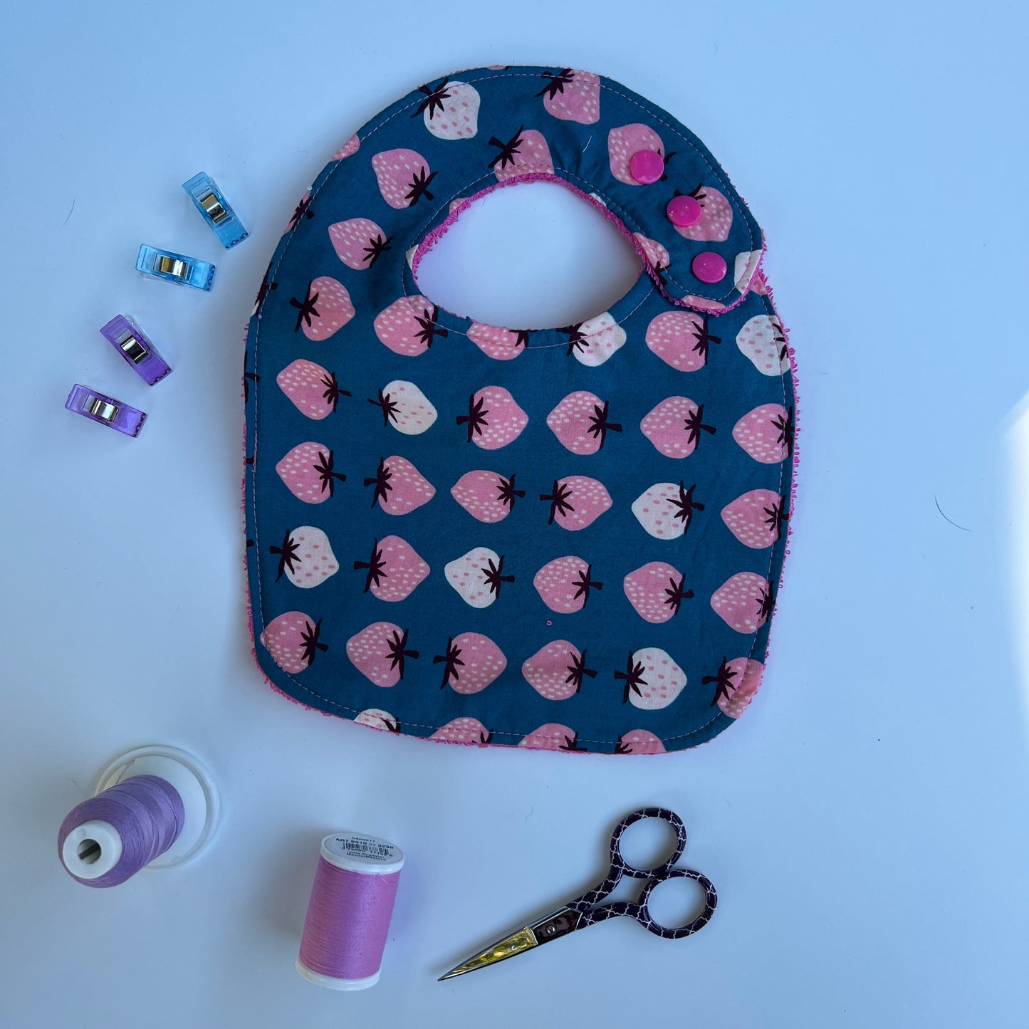 Fabric with strawberry pattern, scissors, spools of thread, and sewing clips on a white background